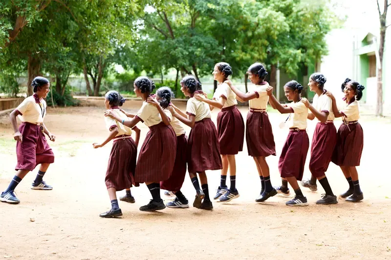 A line of Indiana girls in matching tan and maroon school uniforms hold each others shoulders and playfully skip across a dirt schoolyard. Trees and a school building can be seen in the background.